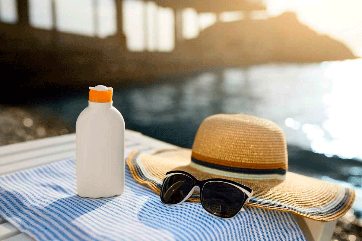 An image of a towel, sun hat, sun glasses and bottle of lotion on a table by a beach.