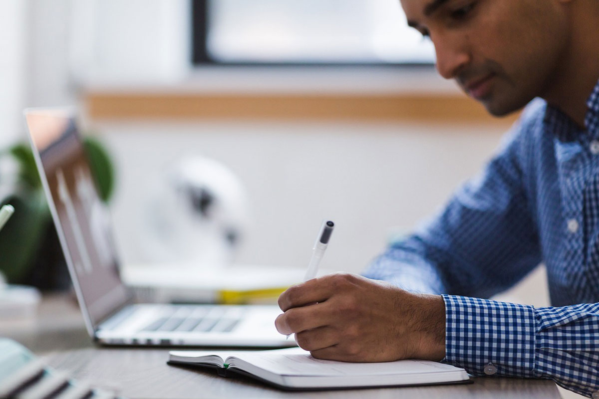 Man looking at a laptop screen writing notes in a notebook